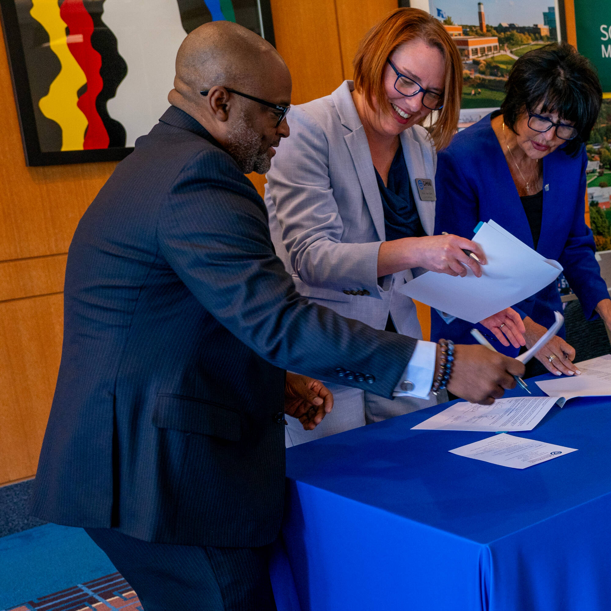 Three people standing at a table with papers, two women in suits and a man in a dark suit.
